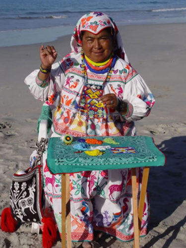 Traditionally dressed woman sitting on chair on the sand sewing with beads.